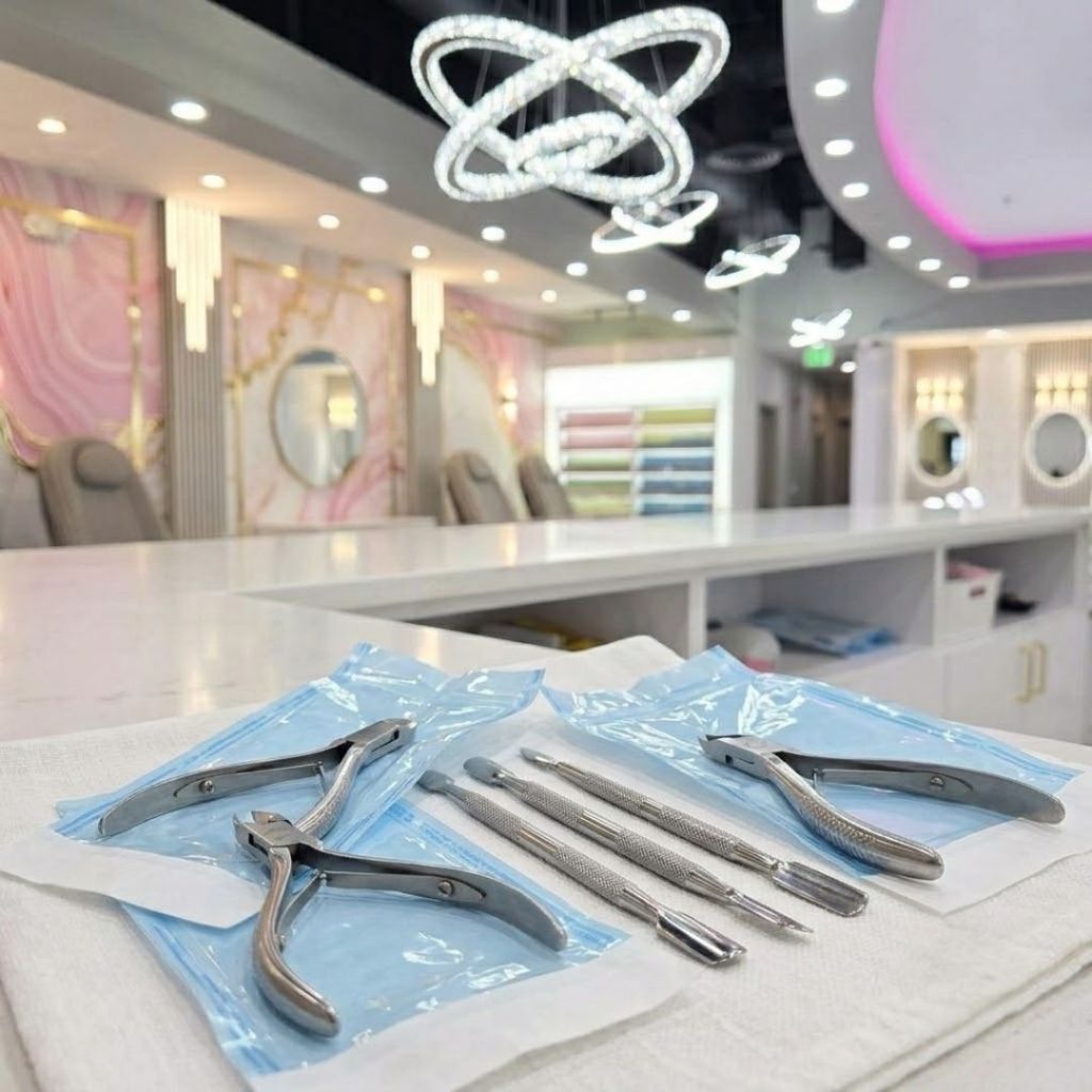 Sterilized metal cuticle tools and pushers inside sealed blue autoclave pouches, resting neatly on a clean white towel on the salon counter under modern crystal chandeliers.